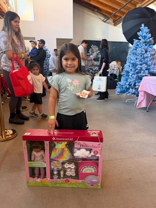 Little girl smiling and displaying the new American Girl doll set she received at the 24LA Holiday Toy Drive sponsored by Omega Law Group.